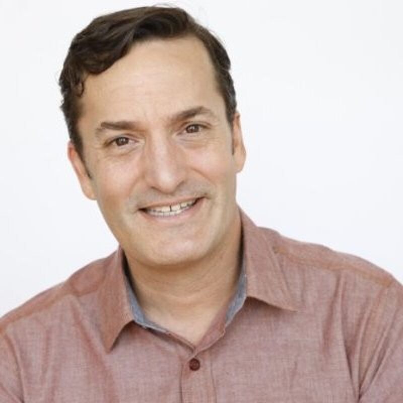 The image shows a middle-aged man with short, dark hair and a friendly smile. He is wearing a reddish-brown button-down shirt. The background is plain white, which makes the man the focal point of the image. The lighting is soft and even, suggesting a professional headshot.